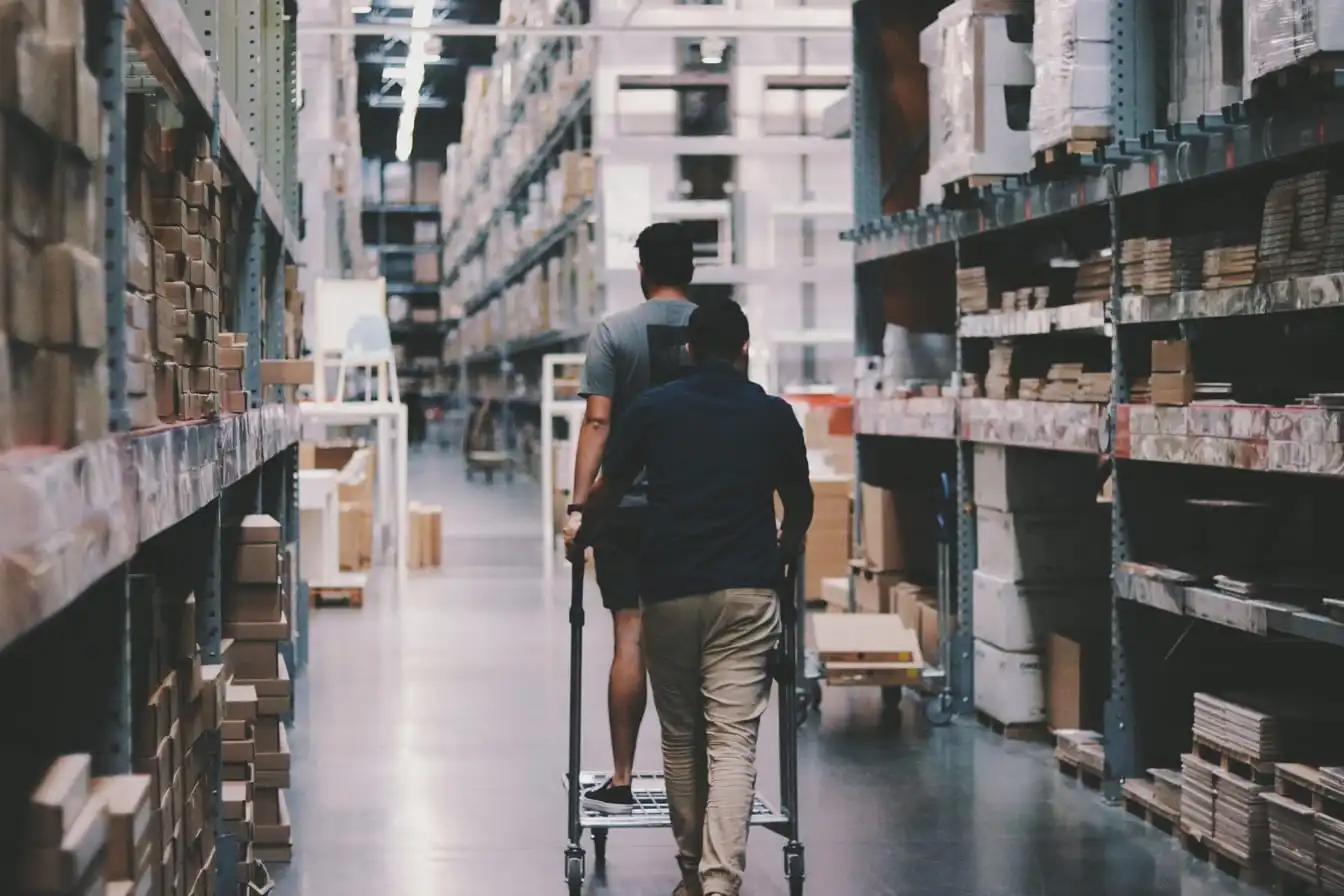 Warehouse shelving stacked with boxes, with two people moving a trolley.